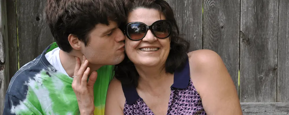 Young adult with autism affectionately kissing his smiling mother outdoors, highlighting love and support during Autism Awareness Month.