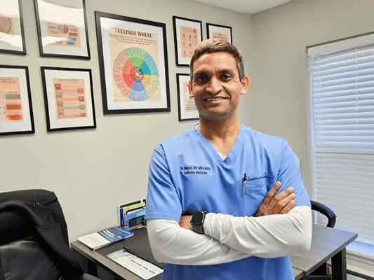 Dr. Nagasasikanth Mopuri is standing in a modern medical office with mental health charts on the wall at a South Jersey clinic supporting reentry healthcare for formerly incarcerated individuals.