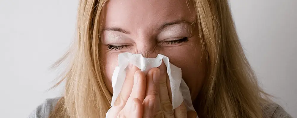 Woman sneezing into a tissue due to seasonal allergies, illustrating common symptoms like a runny nose and congestion from pollen or airborne allergens.